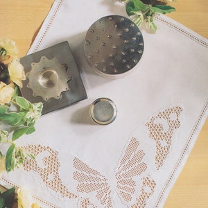 May include: A still-life arrangement featuring a white embroidered linen cloth with a floral design. Silver metal objects, including a round container with holes, a small lid, and a gear-shaped piece, are arranged on the cloth. Yellow roses and greenery add color.