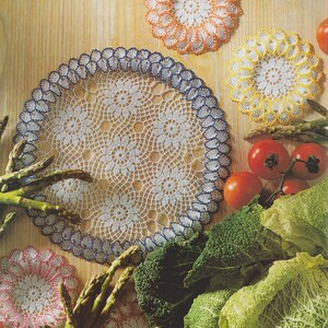 Op de afbeelding: Handgemaakte gehaakte onderzetters in wit, met gekleurde randen in blauw, oranje, geel en roze. De onderzetters zijn gerangschikt op een houten oppervlak met verse groenten, waaronder tomaten, asperges en kool. Het nummer "24" staat linksboven.