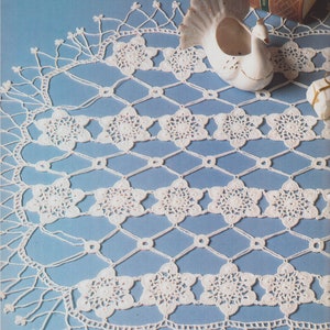 May include: A white, crocheted doily with a floral and geometric pattern. The intricate design features repeating star-shaped flowers connected by a diamond-shaped lattice. The doily is displayed on a light blue surface, with a decorative swan and a book in the background.