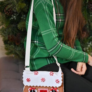 May include: A brown crossbody bag shaped like a gingerbread house with a white scalloped roof featuring red and white peppermint candy accents. The bag has a red door with a green wreath, two windows with white frames, and a white shoulder strap.