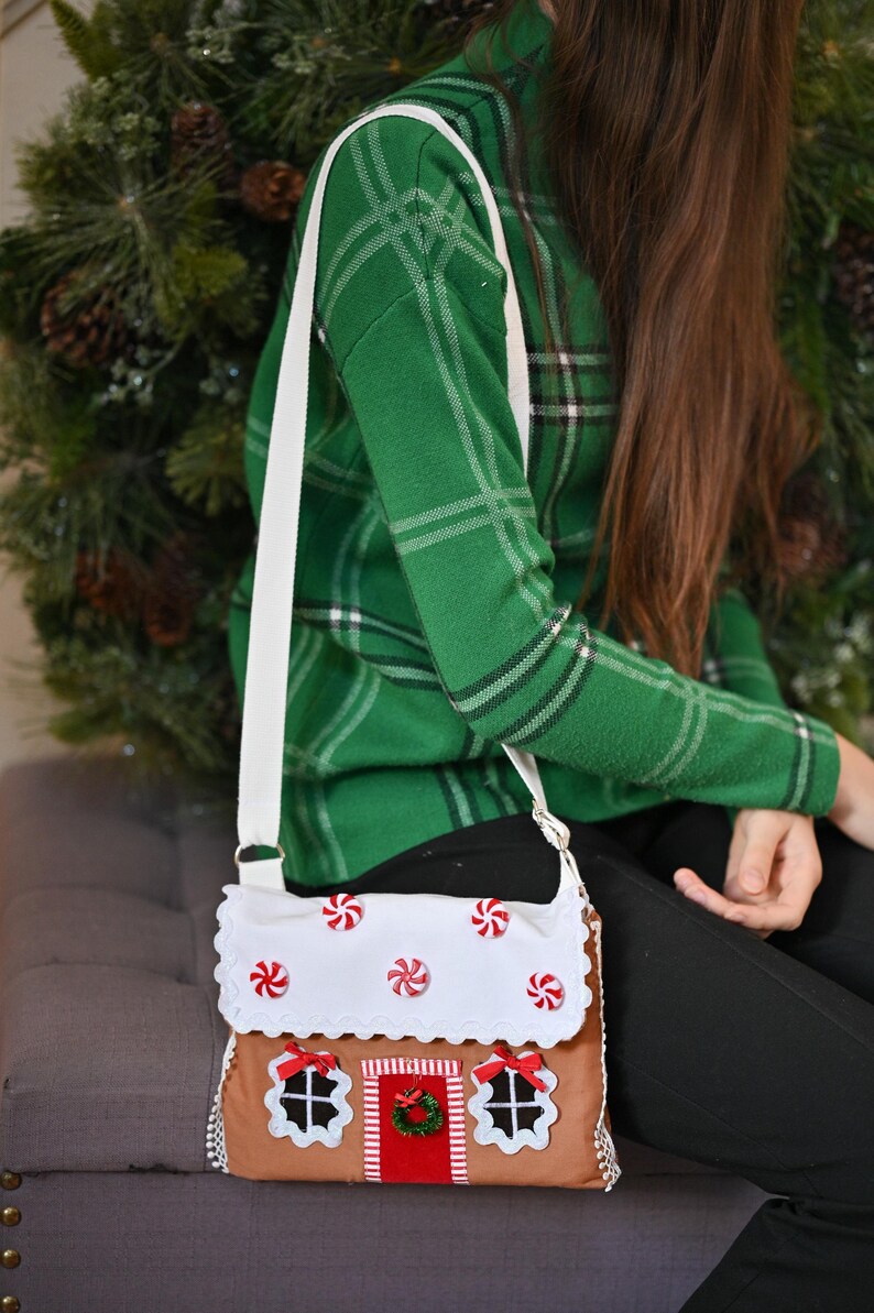 May include: A brown crossbody bag shaped like a gingerbread house with a white scalloped roof featuring red and white peppermint candy accents. The bag has a red door with a green wreath, two windows with white frames, and a white shoulder strap.
