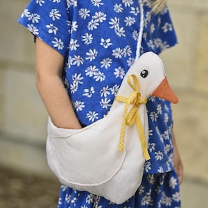 May include: A white fabric goose-shaped shoulder bag with an orange beak, a black eye, and a yellow polka-dot ribbon tied around its neck. The bag features a white cord strap and a deep opening on the back for storage, worn over a blue floral outfit.