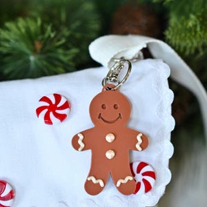 May include: A brown gingerbread man keychain with a smiling face, two white buttons, and white icing details on the arms and legs. The keychain is attached to a white fabric item decorated with small red and white peppermint candy accents.