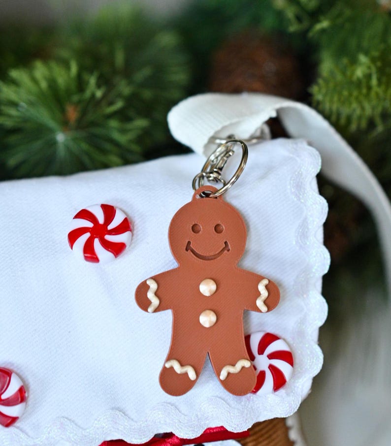 May include: A brown gingerbread man keychain with a smiling face, two white buttons, and white icing details on the arms and legs. The keychain is attached to a white fabric item decorated with small red and white peppermint candy accents.