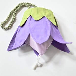 May include: A fabric pouch shaped like a flower with purple petals, a light green top section, and a braided light green cord handle. The interior is a lighter shade of purple, and the opening features a white drawstring with two small white toggles.