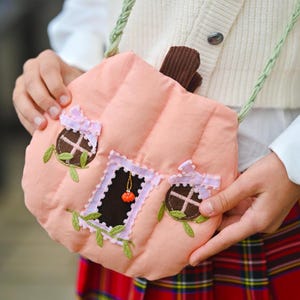 May include: A peach-colored quilted pumpkin-shaped crossbody bag with a brown corduroy stem. The bag features two circular windows and a central door decorated with light pink gingham ribbon trim, green fabric leaves, and a small orange charm.