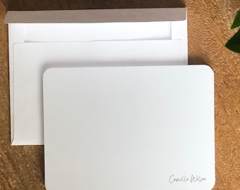 May include: A white rectangular card with rounded corners, placed in a white envelope with a gray flap. The card has a signature in the bottom right corner. The items are on a wooden surface, with green leaves in the background.