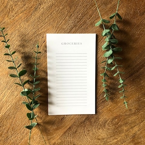 Peut inclure: Un bloc-notes blanc avec le mot "GROCERIES" imprimé en haut. Le bloc-notes a du papier ligné et est posé sur une surface en bois. Il y a deux brins de verdure de chaque côté du bloc-notes.