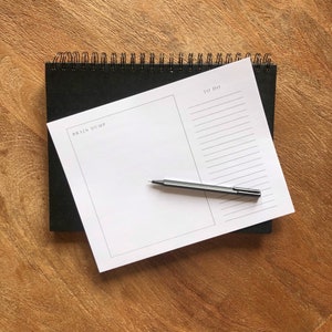May include: A white notepad with a black spiral binding. The notepad is divided into two sections, one labelled "Brain Dump" and the other labelled "To Do". A black pen is lying on the notepad.