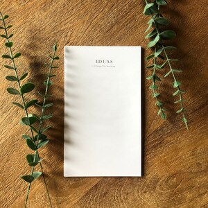 May include: A white notepad with the text "IDEAS I'll forget by morning" printed on the front. The notepad is on a wooden surface with green leafy branches on either side.
