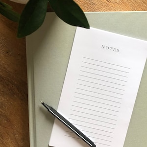May include: A white lined notepad with the word "NOTES" printed at the top. A black pen is lying on the notepad. The notepad is on top of a sage green notebook.