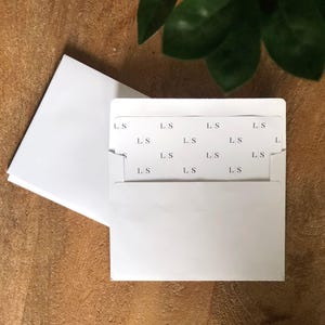 Pode incluir: Envelope branco com um cartão branco dentro. O cartão tem as letras "L.S." repetidas várias vezes.