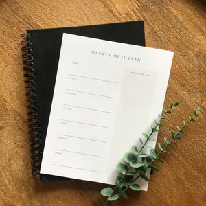 May include: A white weekly meal planner with a grocery list section. The planner is bound with a black spiral binding and is sitting on a wooden surface.
