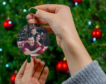 Peut inclure: Une boule de Noël ronde avec une photo d'une famille de trois personnes. La boule est blanche avec un cordon doré et est tenue par une main devant un sapin de Noël.