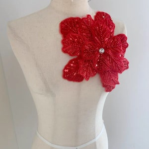 Könnte beinhalten: Eine rote, mit Pailletten besetzte Blumenapplikation mit einem klaren Strassstein in der Mitte. Die Applikation befindet sich auf einer weißen Schaufensterpuppe.