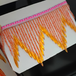 Könnte beinhalten: Nahaufnahme einer dekorativen Franse mit orangefarbenen und gelben Perlen. Die Franse hat einen rosa-weiß gemusterten oberen Rand. Die Perlen sind in vertikalen Linien aufgereiht, mit größeren, tropfenförmigen Perlen unten.