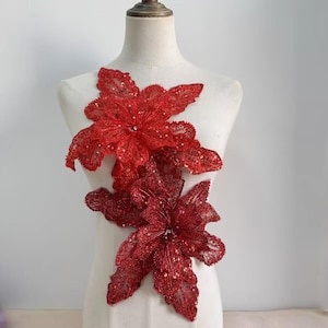 Op de afbeelding: Twee rode, met pailletten versierde bloemenapplicaties. De bloemen hebben meerdere bloemblaadjes en zijn bevestigd aan een mannequin. De applicaties zijn dieprood van kleur en zijn versierd met pailletten, wat een vleugje glans en textuur toevoegt.