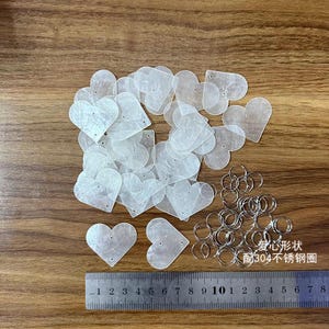 May include: A pile of white heart-shaped plastic pieces with two holes in each piece. There are also a pile of silver metal rings. A ruler is in the bottom of the image with the text "1 2 3 4 5 6 7 8 9 10 1 2 3 4 5 6 7 8" and the text "爱心形状 配304不锈钢圈" in the bottom right corner.