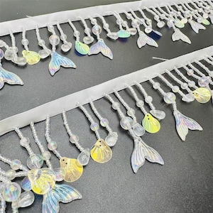 Op de afbeelding: Twee rijen decoratieve sierband met een witte lintbasis. Heldere kralen en iriserende zeemeerminstaartbedels hangen aan het lint. Sommige bedels zijn geel, blauw en groen. De sierband is geschikt voor het verfraaien van kleding of ambachten.