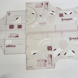 Può includere: Una collezione di strumenti di disegno in plastica trasparente, tra cui righelli, goniometri e modelli. Gli strumenti presentano segni di misurazione, forme geometriche e testo. Gli strumenti sono disposti su una superficie bianca.