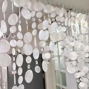 Pode incluir: Uma cortina de contas branca com discos de concha ovais e detalhes de cristal transparente. A cortina pende de uma série de pequenos ganchos de metal, criando um efeito decorativo brilhante. Adequada para portas ou janelas.