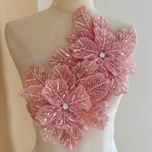 Op de afbeelding: Twee grote roze bloemen van stof met pailletten en kralen. De bloemen hebben een strasssteen in het midden en zijn op elkaar gelegd.