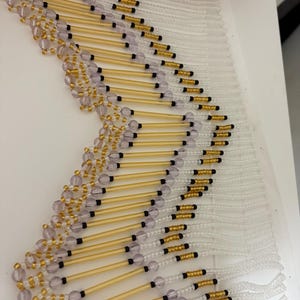 Puede incluir: Un borde de flecos con cuentas decorativas que presenta cuentas tubulares largas de tono dorado, cuentas redondas translúcidas de color violeta claro y pequeñas cuentas negras. El diseño tiene un patrón de chevrón con hilos de cuentas blancas.
