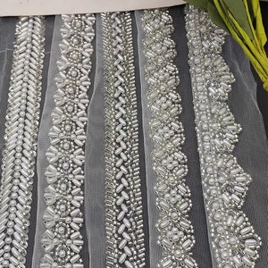 Puede incluir: Cinco diferentes galones de cuentas en blanco y plateado sobre una tela blanca transparente. Los galones son todos de diseños diferentes y están dispuestos en una fila.