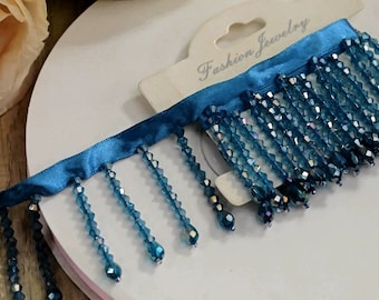 May include: Teal blue beaded fringe trim with a satin ribbon backing. The trim is labelled "Fashion Jewelry".