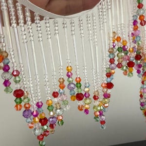 Puede incluir: Un fleco decorativo de cuentas con una cinta blanca. El fleco presenta cuentas claras y facetadas y cuentas coloridas en tonos de rojo, naranja, amarillo, verde, morado y rosa. Las cuentas están ensartadas en cordones transparentes.