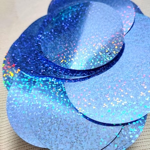May include: A collection of iridescent blue sequins, each circular with a small hole. The sequins have a glittery, reflective surface, creating a shimmering effect. The sequins are scattered and overlapping, with a range of blue hues.