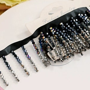 Könnte beinhalten: Schwarzes Bandbesatz mit einem Fransenbesatz aus schimmernden schwarzen und silbernen Perlen. Der Besatz ist mit "Fashion Jewelry" beschriftet.
