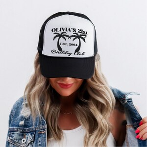 May include: A black and white trucker hat with the text "Olivia's 21st Birthday Club" and palm trees. The text "EST. 2004" is also on the hat.