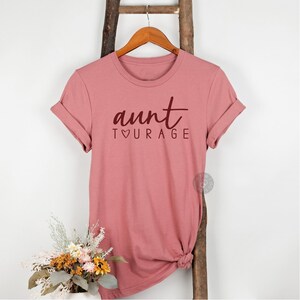 May include: A dusty rose short-sleeved t-shirt featuring the dark maroon text "aunt" in a cursive script above the word "TOURAGE" in a clean sans-serif font, with a small heart symbol replacing the letter "O" in the word "TOURAGE".