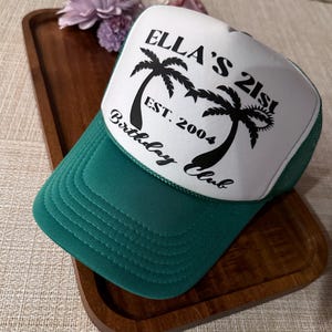 May include: A green and white trucker hat with the text "Ella's 21st Birthday Club Est. 2004" and palm trees printed on the front.