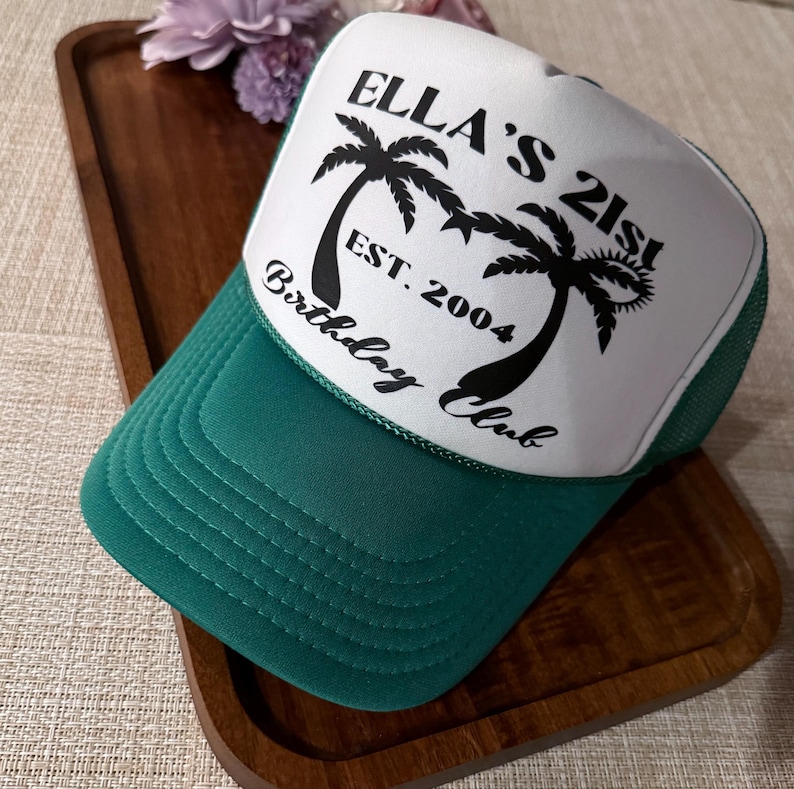 May include: A green and white trucker hat with the text "Ella's 21st Birthday Club Est. 2004" and palm trees printed on the front.