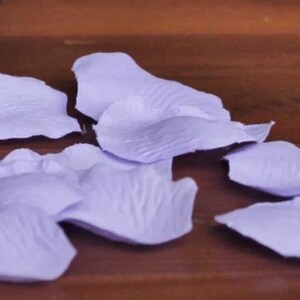 May include: A scattering of lavender silk rose petals on a wooden surface. The petals are soft and delicate, with a slightly textured surface.