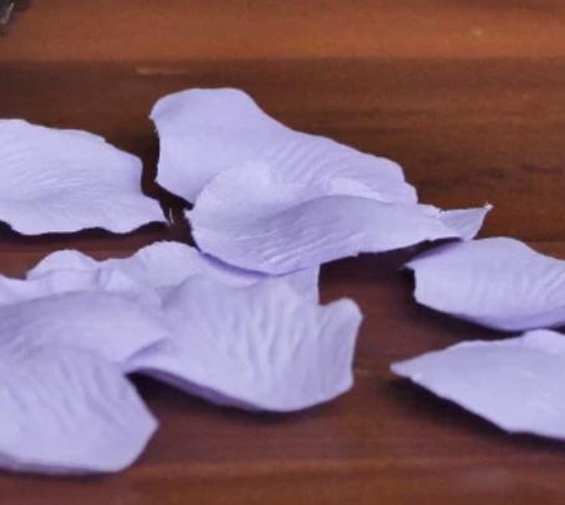 May include: A scattering of lavender silk rose petals on a wooden surface. The petals are soft and delicate, with a slightly textured surface.