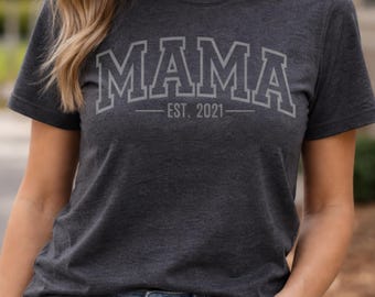 May include: Dark gray t-shirt with the word "MAMA" in large, outlined letters. Below the word "MAMA" is the text "EST. 2021". The shirt has short sleeves and a crew neck.