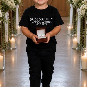 May include: A black t-shirt with the text "BRIDE SECURITY JACKSON WEDDING 06.21.2028" is worn by a child. The child is holding a small, open, wooden ring box with a ring inside. The child is wearing black pants.