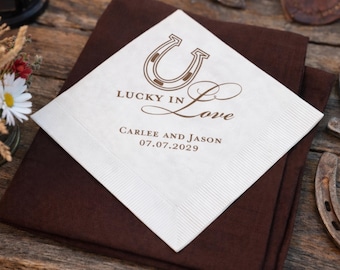 May include: A white square napkin with a brown horseshoe and the words "Lucky in Love" printed on it. Below the text are the names "Carlee and Jason" and the date "07.07.2029". The napkin is on top of a brown napkin.