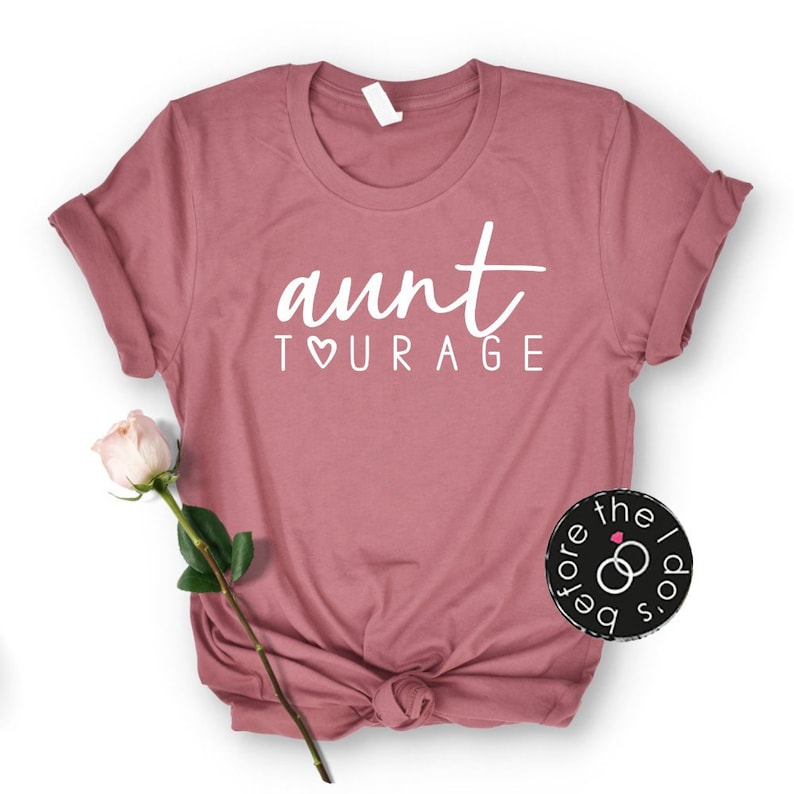 May include: A mauve short-sleeved t-shirt featuring the white text "aunt TOURAGE" with a heart replacing the letter O. A single pale pink rose with a green stem and leaves is positioned to the left of the shirt, and a circular logo is on the right.