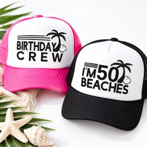 May include: Two trucker hats with white front panels and mesh backs. The hot pink hat features black text reading BIRTHDAY CREW with a palm tree and seashell graphic. The black hat features black text reading I'M 50 BEACHES with a palm tree and seashell.