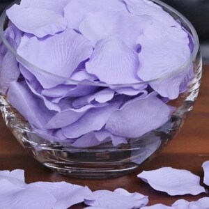 May include: A glass bowl filled with lavender-colored rose petals. The petals are scattered on a wooden surface.