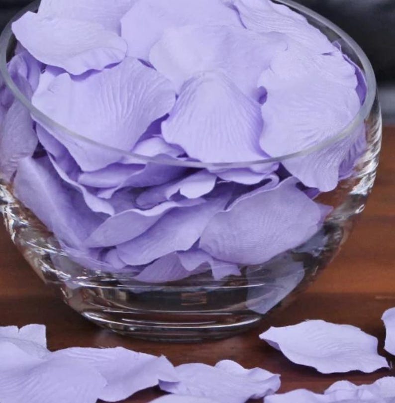 May include: A glass bowl filled with lavender-colored rose petals. The petals are scattered on a wooden surface.
