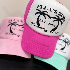 May include: Three trucker hats with pink, white and light blue mesh backs. The front of the hats are white with black palm trees and the text "Ella's 21st Birthday Girl Est. 2004".