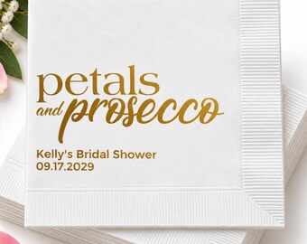 May include: A square white paper napkin with a textured border. The text "petals and prosecco" is printed in gold, followed by "Kelly's Bridal Shower" and the date "09.17.2029" in a smaller, simple gold font centered on the napkin surface.