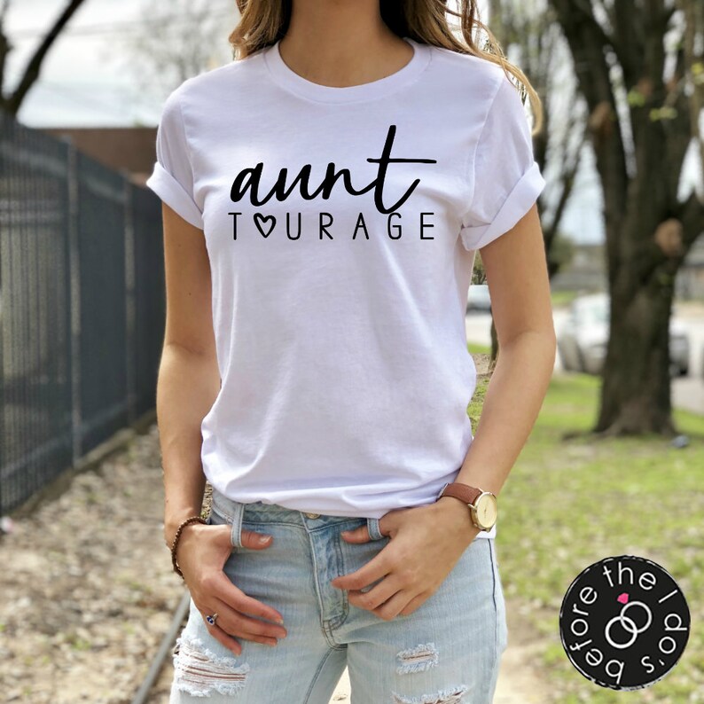 May include: A white short-sleeved t-shirt with a crew neckline and rolled sleeves. The front of the shirt features the black text "aunt" in a cursive script above the word "TOURAGE" in a sans-serif font, with a heart replacing the letter O.