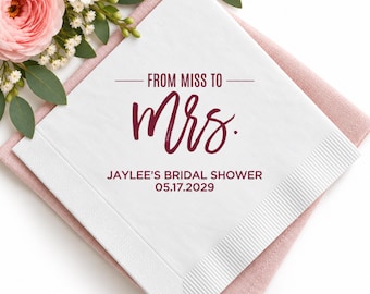 May include: A white square paper napkin featuring dark red text that reads FROM MISS TO Mrs. JAYLEE'S BRIDAL SHOWER 05.17.2029. The napkin has a textured border and is placed on a pink fabric surface next to pink flowers and green leaves.