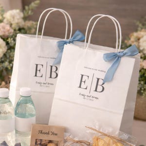 May include: Two white paper gift bags with handles, each adorned with a light blue ribbon. The bags feature the initials "EB" and the text "Emily and Brunn 10/11/2025" printed in black. The bags are part of a wedding favor set.